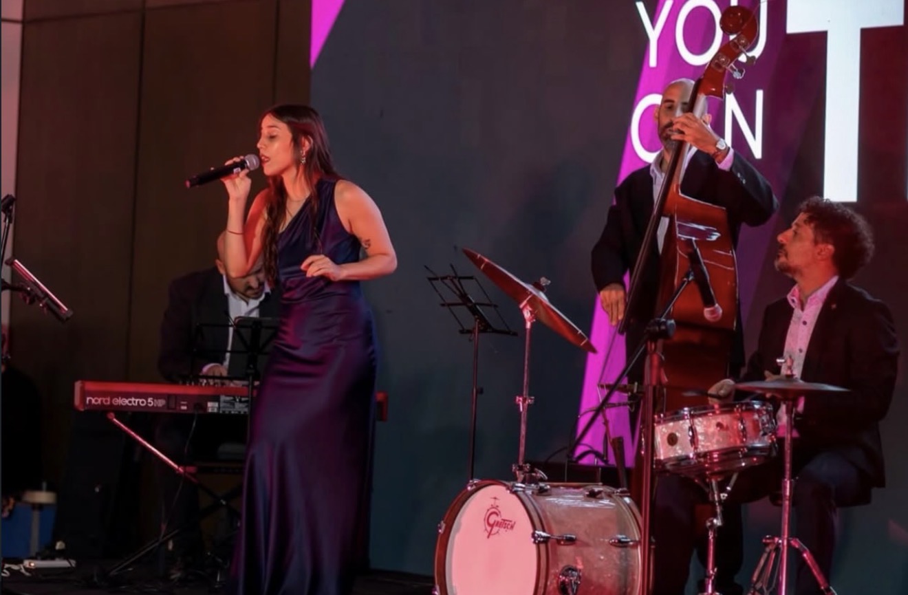 Cantante con micrófono, teclado, contrabajo y batería en escenario interior: Harmony Buenos Aires.